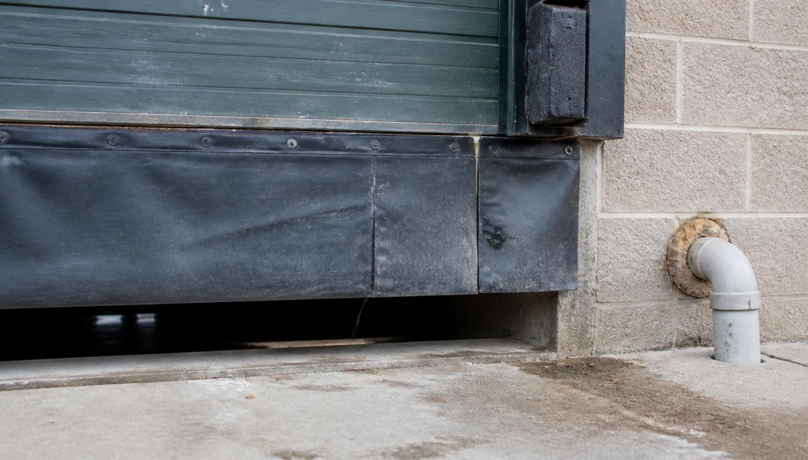 Gap beneath a commercial loading dock door with a worn rubber sweep and an unsealed utility pipe penetration in the block wall