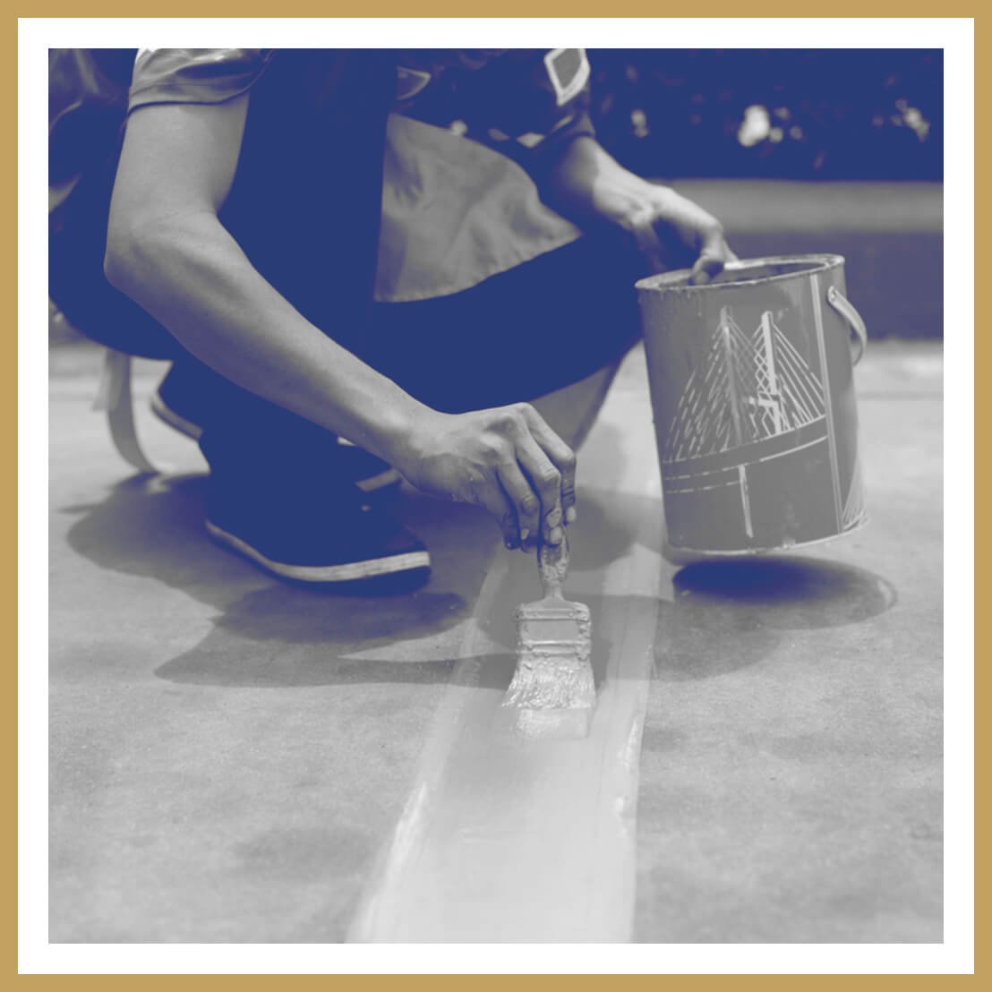 A person crouches and paints a white line on the ground with a brush, holding a paint can in their other hand. The image has a blue and white filter and a beige border.