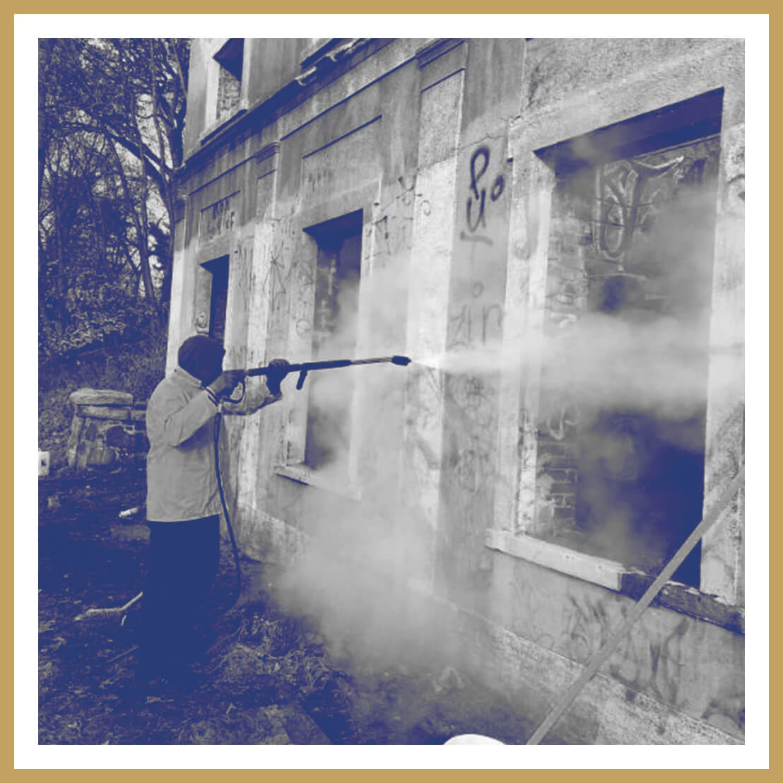 A person uses a power washer for graffiti removal on the exterior wall of an old, abandoned building with broken windows. Thick clouds of water spray cover parts of the wall.