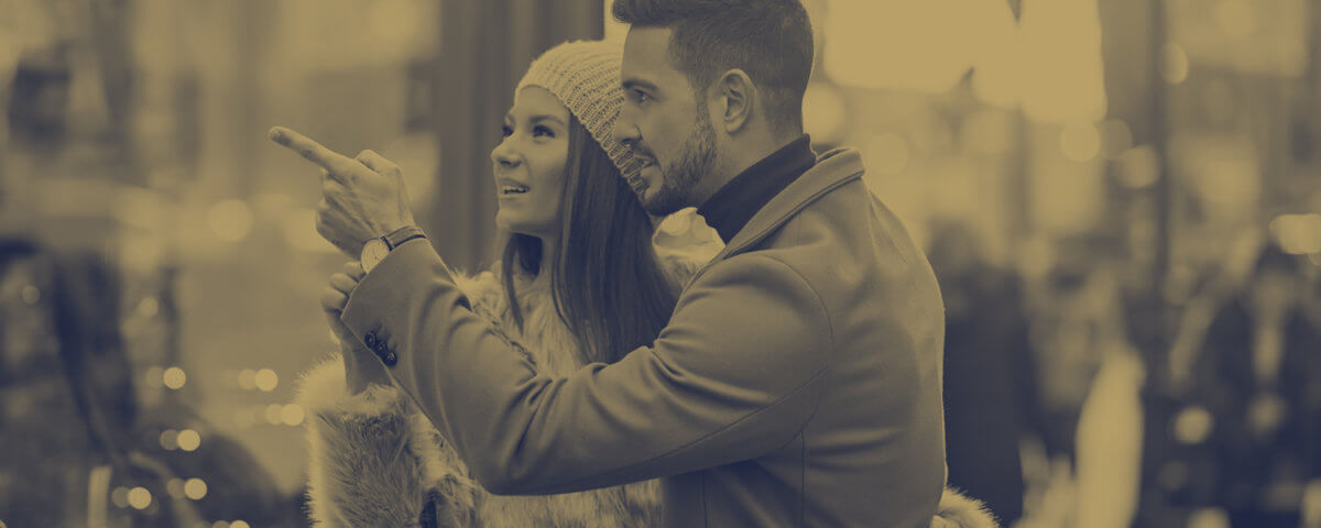 A couple dressed in winter clothing stands close together, smiling as they look and point at a festive storefront window display on a city street. The image has a warm, sepia tone and the Nationwide Bank Insurance logo is visible.