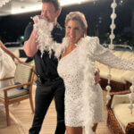 A smiling woman in a sparkling white dress and feather boa poses on a yacht with a man in a black outfit who playfully hides behind the boa. The setting features string decorations and outdoor seating.