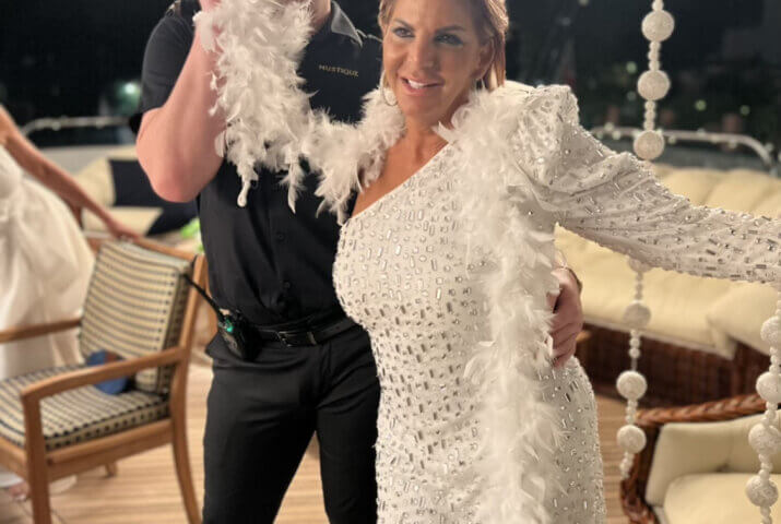 A smiling woman in a sparkling white dress and feather boa poses on a yacht with a man in a black outfit who playfully hides behind the boa. The setting features string decorations and outdoor seating.