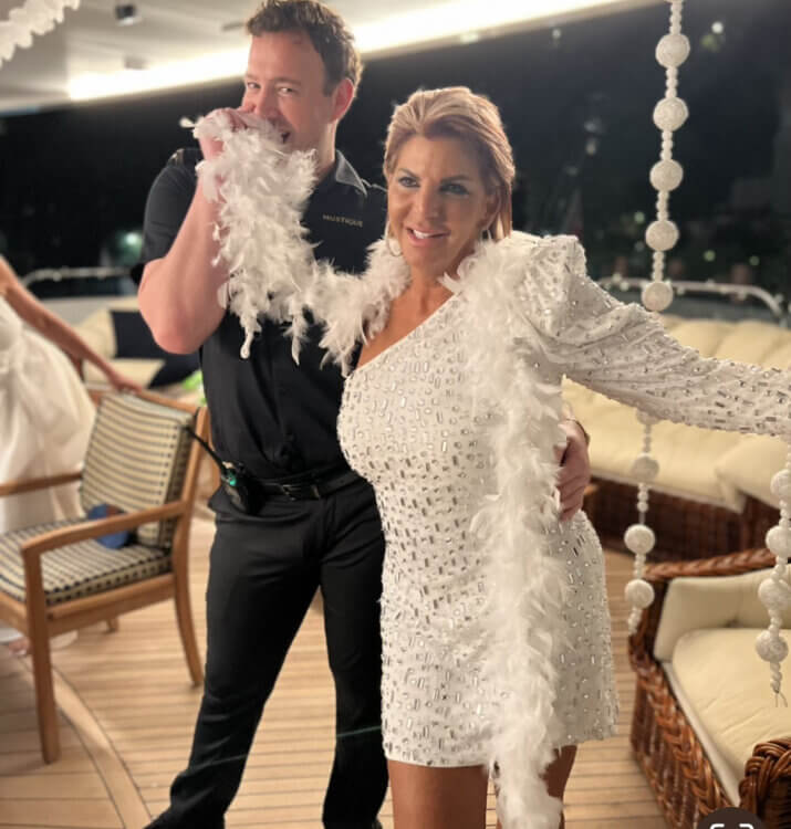 A smiling woman in a sparkling white dress and feather boa poses on a yacht with a man in a black outfit who playfully hides behind the boa. The setting features string decorations and outdoor seating.