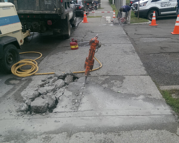 A jackhammer stands upright in the middle of a broken concrete sidewalk, surrounded by rubble. Construction vehicles, equipment, and orange traffic cones are visible, with workers in the background.