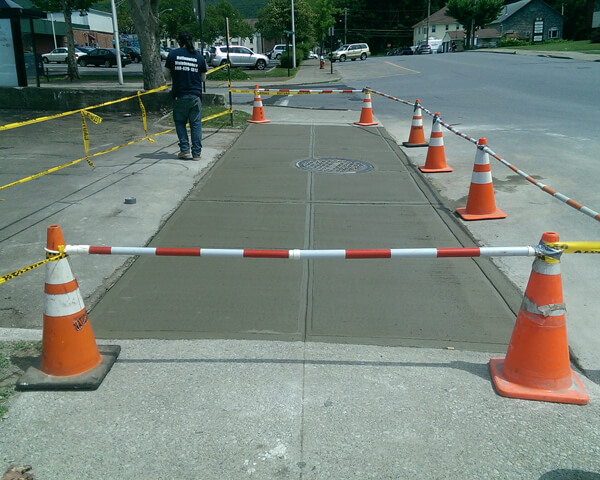 Freshly poured concrete sidewalk section, blocked off with orange cones, caution tape, and a striped barrier. A person stands nearby, wearing work clothes. Cars and buildings are visible in the background.