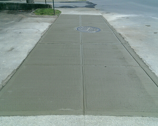 Freshly poured concrete sidewalk with clear rectangular sections and a round metal manhole cover near the top, bordered by asphalt and patches of grass.