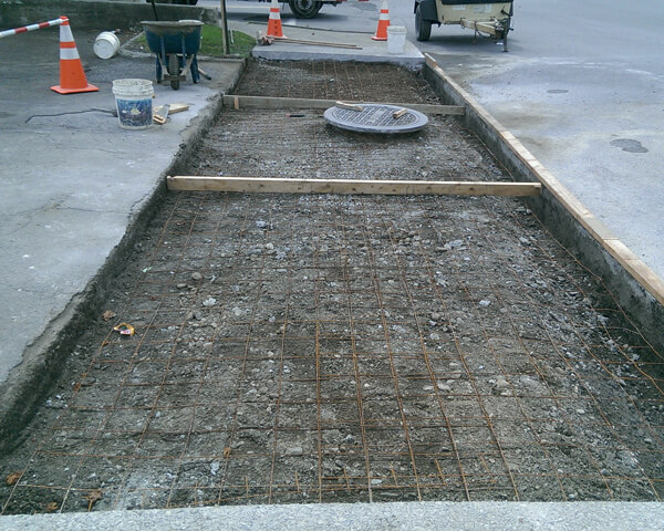 A section of sidewalk under construction with exposed rebar grid, loose dirt, traffic cones, a bucket, and a wheelbarrow nearby. A manhole cover is raised above ground level in the middle of the area.