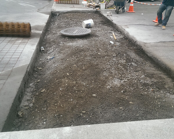 A section of a city sidewalk under construction with exposed soil, a raised manhole cover, construction materials, and orange safety cones visible in the background.