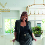 A woman in a black outfit stands in a modern kitchen with white cabinets and gold accents. She is smiling and leaning on a counter. The logo Nationwide Maintenance appears in the top left corner.