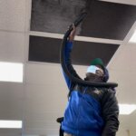 A person wearing a mask and blue jacket uses a vacuum hose to clean a vent on a ceiling tile in a brightly lit indoor space.
