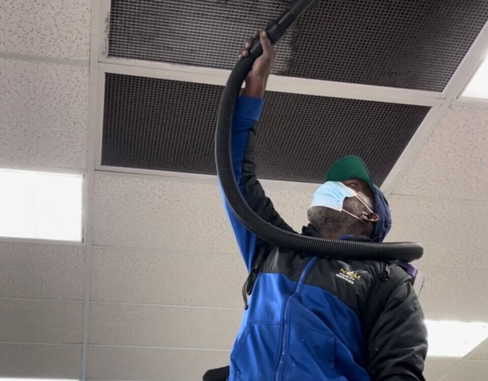 A person wearing a mask and blue jacket uses a vacuum hose to clean a vent on a ceiling tile in a brightly lit indoor space.