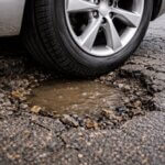 A car tire is partially submerged in a large, water-filled pothole on a damaged asphalt road.