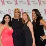 Four women in formal dresses stand together, smiling, in front of a step-and-repeat banner with pink breast cancer ribbons and words like “Building Hope,” “Faith,” and “A Cure.”.