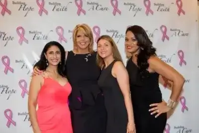 Four women in formal dresses stand together, smiling, in front of a step-and-repeat banner with pink breast cancer ribbons and words like “Building Hope,” “Faith,” and “A Cure.”.