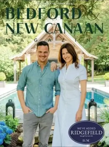 A man and a woman stand smiling in front of a pool and wooden gazebo, surrounded by lush greenery, on the cover of Bedford & New Canaan Magazine, September/October 2023 issue.