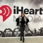 A woman in a black outfit with a fur collar stands smiling in front of an iHeartMedia wall featuring a large red logo and silhouettes of a cheering concert crowd.