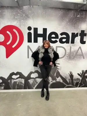 A woman in a black outfit with a fur collar stands smiling in front of an iHeartMedia wall featuring a large red logo and silhouettes of a cheering concert crowd.
