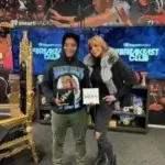 Two people stand and smile in a radio studio, holding signs. Behind them is a colorful mural of faces and “The Breakfast Club” logo on TV screens. The studio features a gold ornate chair and shelves with books and decorations.