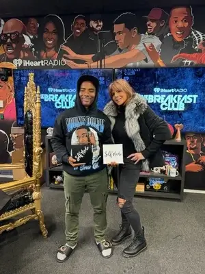 Two people stand and smile in a radio studio, holding signs. Behind them is a colorful mural of faces and “The Breakfast Club” logo on TV screens. The studio features a gold ornate chair and shelves with books and decorations.