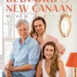 Three smiling adults, two women and one man, pose together indoors in front of a window on the cover of Bedford & New Canaan Magazine, July/August 2023 issue. The setting is bright and warmly decorated.