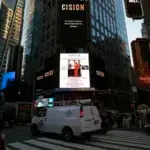 A busy Times Square intersection at dusk, with cars and pedestrians. A white van is in the foreground. Digital billboards, including one displaying POWER: Honore, Maura McMahon Schanzer, light up the tall buildings.