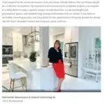 A woman stands and smiles in a modern white kitchen with gold accents, plants, and stylish decor. She wears a black jacket and red skirt, resting her hand on the white marble kitchen island.