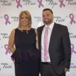 A woman and a man dressed formally stand together and smile in front of a step-and-repeat banner with pink breast cancer ribbons and text reading “Building Hope, Faith, A Cure.”.