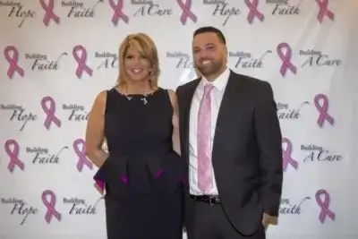 A woman and a man dressed formally stand together and smile in front of a step-and-repeat banner with pink breast cancer ribbons and text reading “Building Hope, Faith, A Cure.”.