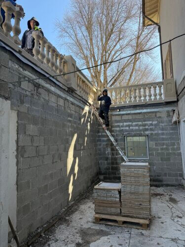 Two workers stand on a precarious wooden plank placed diagonally between a stack of tiles and a second-story balcony of an unfinished building, with bare trees visible in the background.