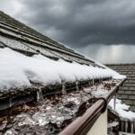 Close-up of a house roof with snow melting into an ice-filled gutter. Dark storm clouds fill the sky in the background, and some raindrops are visible, indicating a transition from snow to rain.