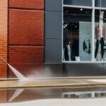 A man wearing overalls and yellow gloves uses a pressure washer to clean a sidewalk outside a building with brick and glass walls, with mannequins visible in the store window.