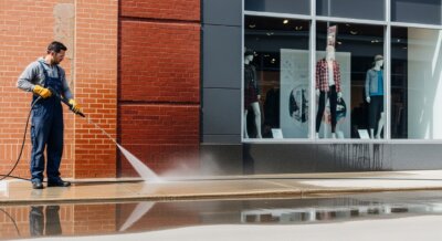 A man wearing overalls and yellow gloves uses a pressure washer to clean a sidewalk outside a building with brick and glass walls, with mannequins visible in the store window.